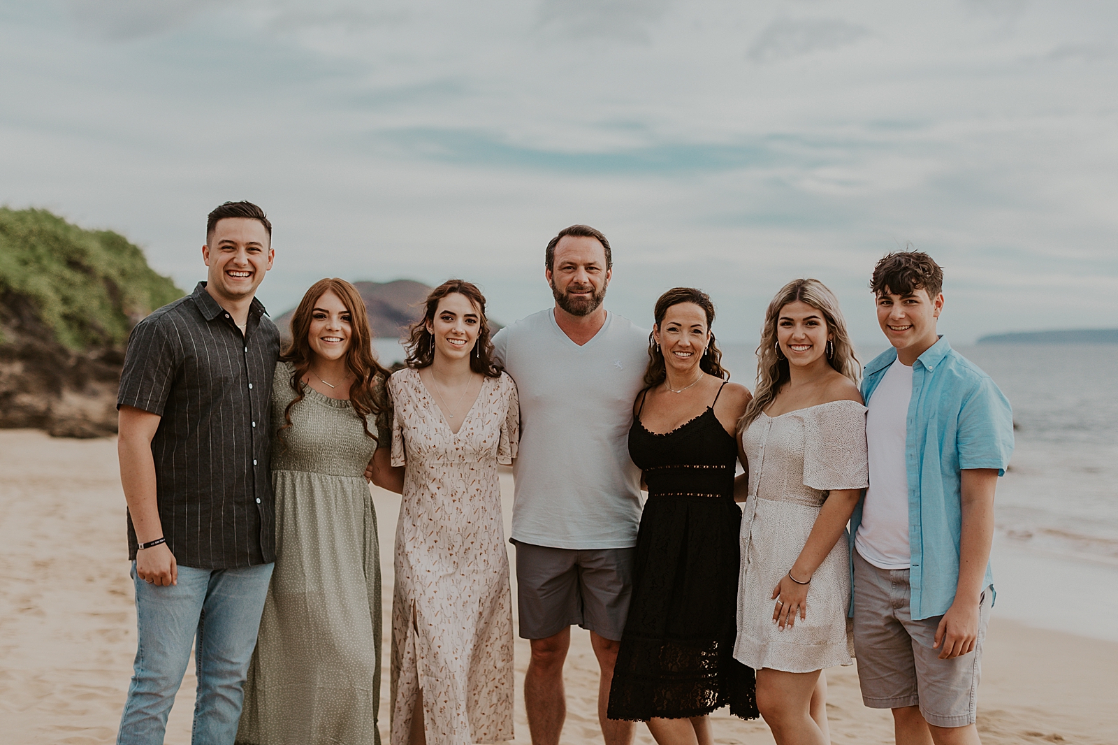 Entire grown up family arms wrapped around each other on the beach in front of the ocean