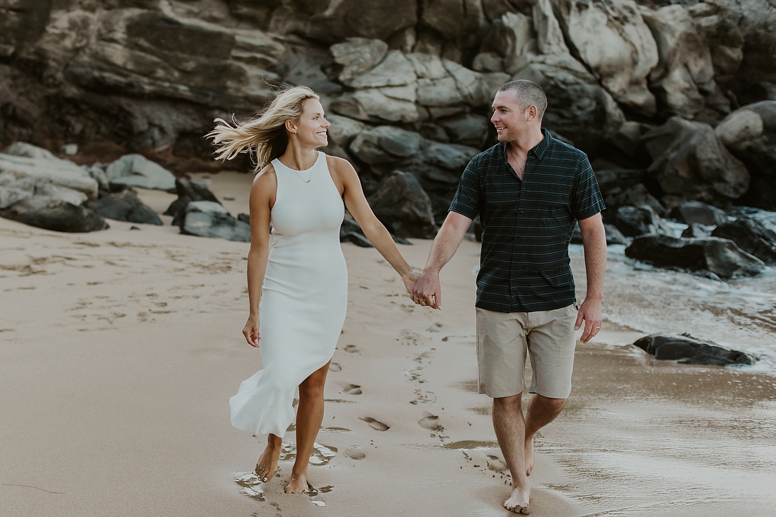 Couple holding hands and walking on the beach together with rocky cliff behind them