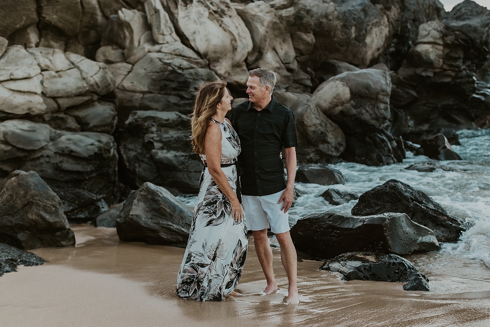 Old couple standing by rocky cliffside on the sand and looking at each other