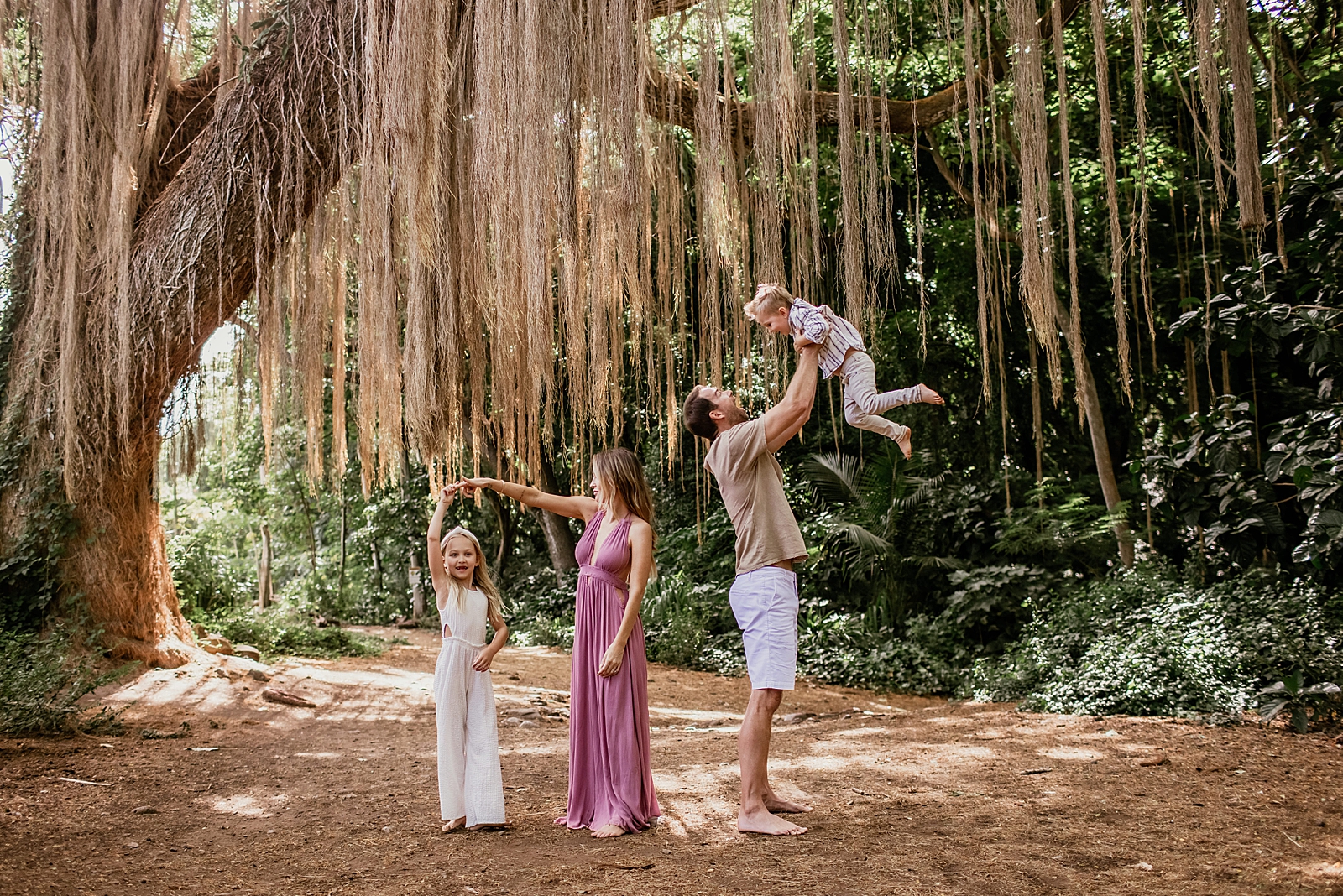Dad throwing up son in the air and daughter twirling by Mother's hand while they are under tree