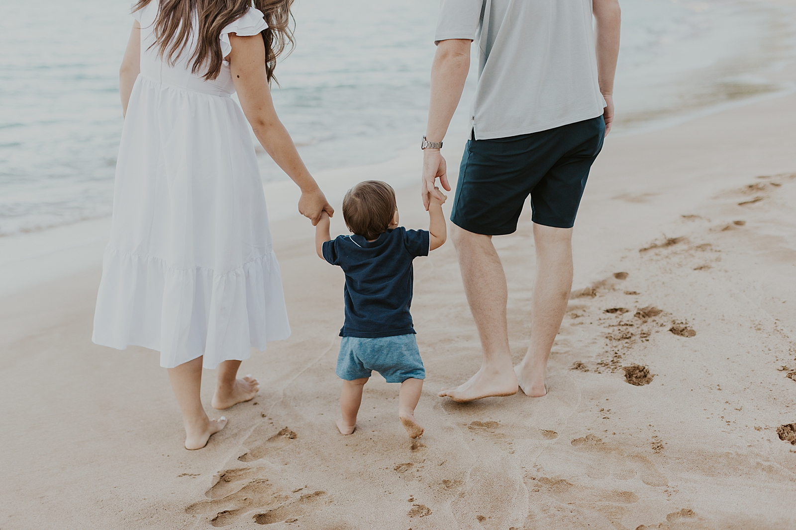Parents holding toddler's sons' hands and walking on the sand of the beach together