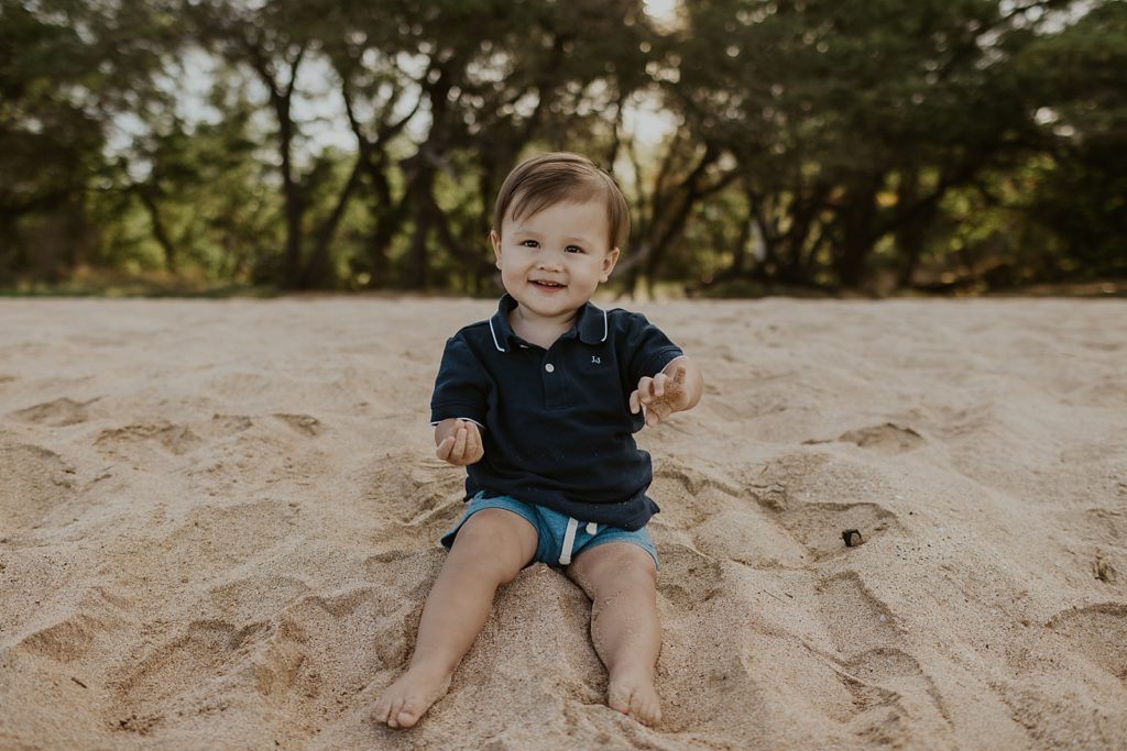 Toddler son sitting on sand of the beach with trees behind him