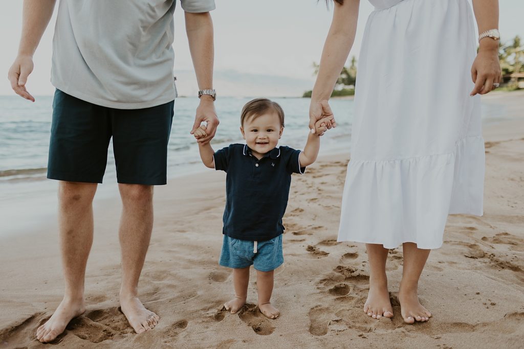 Closeup of son holding parents hands on the sand of the beach with the ocean behind them